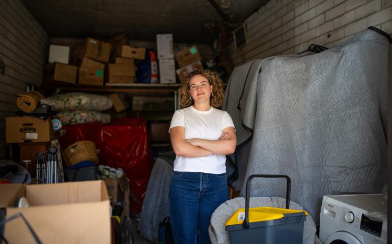 Sarah, wears a white tshirt and jeans standing among her boxed up belongings inside a garage